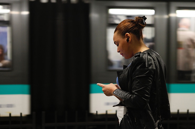 女性が地下鉄のホームでワイヤレスイヤホンを装着してスマホを操作している画像